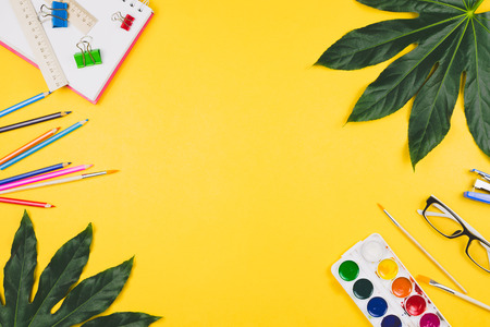 Business flatlay with tropical leaves and business and artistic accessories: colored pencils, rulers, notebook, clips, watercolors etc. Top view, yellow background. Concept of study or creative work.の写真素材
