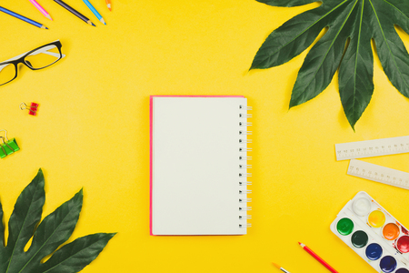 Business flatlay with tropical leaves and business and artistic accessories: colored pencils, rulers, notebook, clips, watercolors etc. Top view, yellow background. Concept of study or creative work.の写真素材