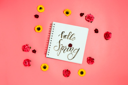 Notebook with white page with handletter "Hello Spring" and various artificial flowers around. Flatlay, pink background.の写真素材