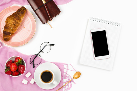 Beautiful flatlay arrangement with croissant, cup of coffee, fresh strawberries, smartphone with black copyspace and other business accessories: concept of busy morning breakfast, white background.の写真素材
