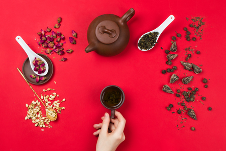Flatlay frame arrangement with Chinese green tea leaves, rose buds, jasmine flowers, tea pot and woman's hands holding tea cup. Red backgroundの写真素材
