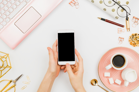 Stylish flatlay frame arrangement with woman's hands holding white smartphone with black copyspace, pink laptop, coffee, planner and other accessories. Feminine business mockupの写真素材