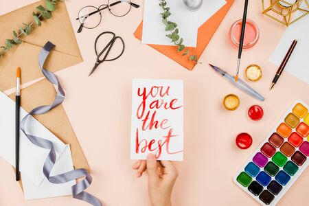 Stylish flatlay with art supplies, envelopes, brushes, watercolors, glasses, pen and woman's hand holding a handmade card with handlettered sign "You are the best". Pastel backgroundの写真素材