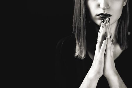 Portrait of young adult white woman wearing dark plum lipstick and holding her hands with dark manicure in a pray like gesture near her face. Selective focus. Black and white, dramatic photo.の写真素材