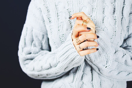 Closeup of a woman's hands joined together in a gray sweater with golden rings and dark manicure.