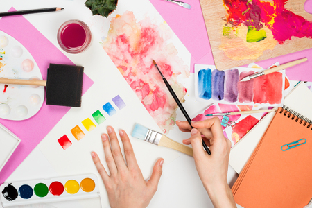 Flatlay with woman's hands holding brush, paletts, pencils, watercolors, colored paper and other stationary supplies, artist or designer workplaceの写真素材