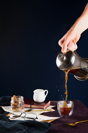 Closeup of man's hand pouring black coffee from french press into a cup on a wooden table with glasses, milk holder, cane sugar and notebook, selective focusの写真素材
