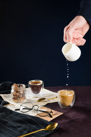 Closeup of men' hand pouring plant milk into cup of coffee on a table with vintage notes, towel, glasses, spoon and jar with sugar, selective focusの写真素材