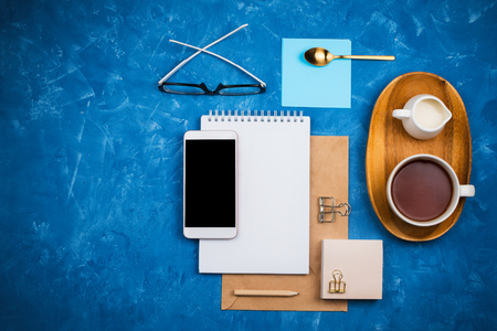 Stylish business flatlay mockup with notebook, glasses, pencil, milk holder and tea on wooden tray and smartphone with black copyspace knolled together on blue cement backgroundの写真素材