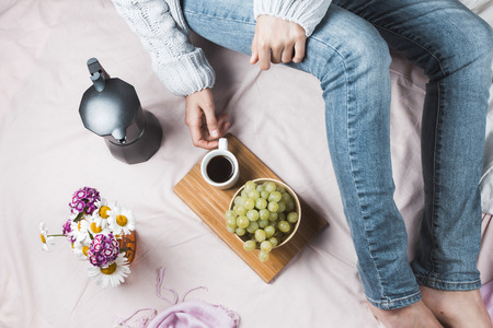 Cozy flatlay of woman in jeans and soft sweater sitting in her bed with cup of coffee and green...