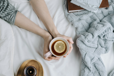 Cozy flatlay of woman's hand holding lemon tea in bed with white sheets and blanket, knitted sweater and candleの写真素材