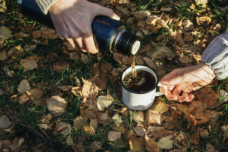 Closeup of woman's and man's hands holding metal cup and flask bottle and pouring tea on the grass in the woods outdoors, selective focusの写真素材