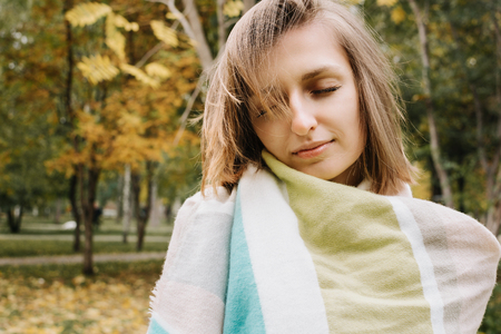 Lifestyle portrait of young adult woman wrapped in scarf with her eyes closed in forest or park, selective focusの写真素材