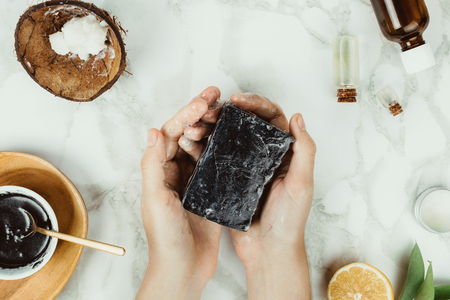 Flatlay of woman's hands holding homemade charcoal soap with its ingredients on the side, natural home beauty conceptの写真素材
