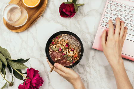 Flatlay of woman's hands holding smoothie bowl with laptop, peonies and water on marble tableの写真素材