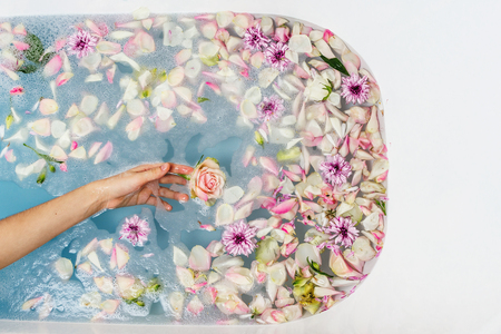 Top view of bath filled with blue bubble water, flowers and petals with woman's hand, spa or...