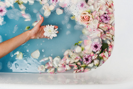 Top view of bath filled with blue bubble water and petals with woman's hand holding flower, spa or...