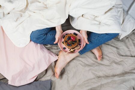 Top view of woman in jeans and white shirt in her bed in a blanket holding vegan whole-wheat pancakes with berries, selective focusの写真素材