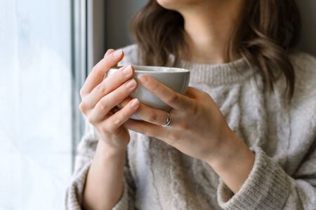 Closeup of woman's hand holding cup with latte in front of the window, shallow selective focus