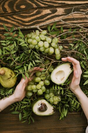 Flat lay of woman's hands holding grapes, pears and avocados with pistachio branches on wooden tableの写真素材