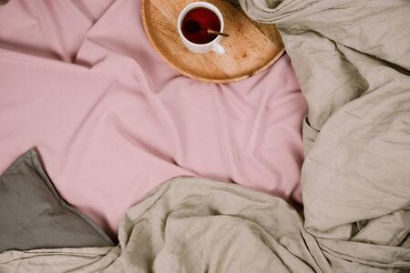 Cozy flatlay of woman's bed with pink sheets and linen grey blanket with tray and tea cup, selective...
