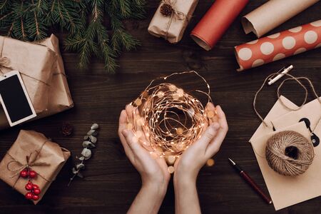 Holiday flatlay arrangement of wrapped presents and woman's hands holding Christmas lights on dark wooden backgroundの写真素材
