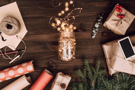Holiday flatlay arrangement of wrapped presents and jar with Christmas lights on dark wooden...