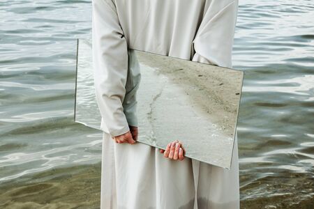 Closeup of woman standind on a shore in blue dress holding a mirror with beach reflecting in it, selectiveの写真素材