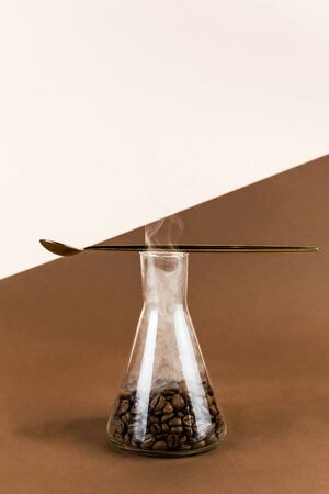 Conical flask with coffee beans on brown and light background with golden spoon and smoke on top, selective focusの写真素材