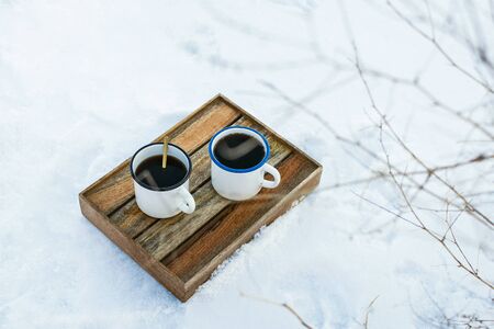 Two enamel mugs with hot drink on wooden box in the snowy field, selective focus