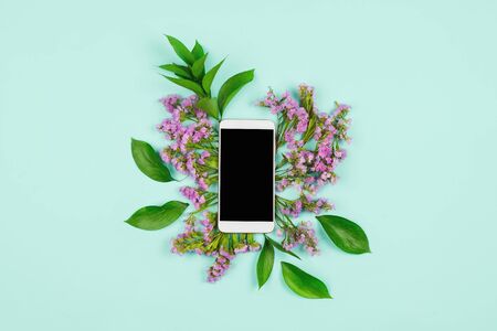 White smartphone with black copyspace screen with leaves and pink wildflowers around. Blue backgroundの写真素材