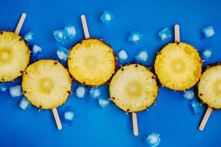 Flatlay of cut pineapples on wooden sticks with ice cubes on blue background