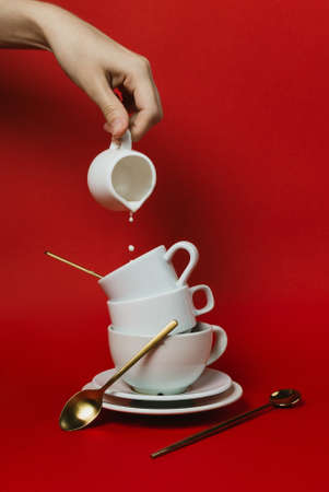 Female hand holding creamer jar and pouring into stack of white coffee cups on redの写真素材