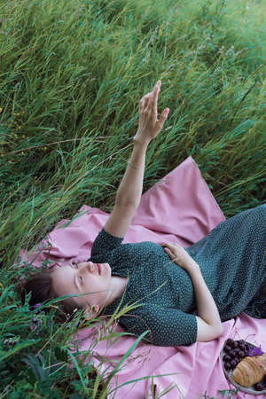 Young adult female in green dress lying on pink blanket and pointing her hand to the sky outdoorsの写真素材