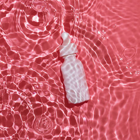 Flatlay of white bottle with serum lying in pink rippling water