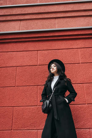 Young asian girl in dark coat and hat standing in front of red wallの写真素材