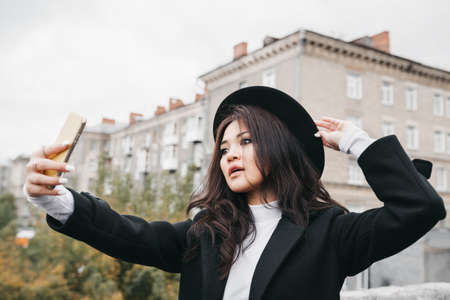 Young asian female in dark coat and hat taking a selfie outdoorsの写真素材