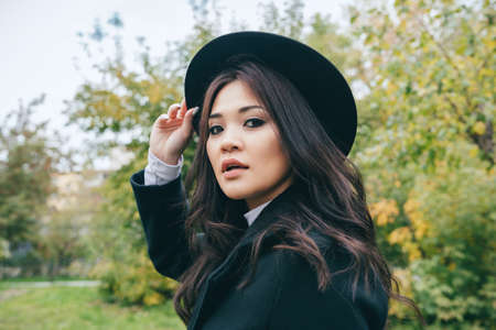Beautiful portrait of young adult asian female in dark coat and hat outdoorsの写真素材