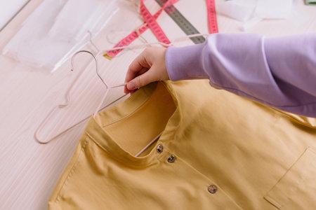 Top view of hands of female seamstress holding finished yellow shirt on a hangerの写真素材