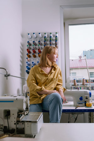 Young female in yellow shirt sitting in her sewing workshop in front of the colorful threads and sewing machines, selective focusの写真素材