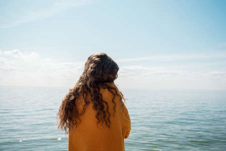 Back shot of female standing on a beach in front of the water in a yellow sweater,の写真素材
