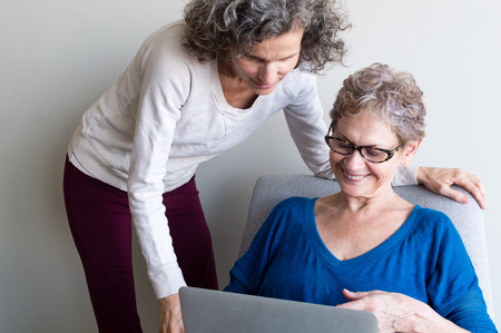 Middle aged woman in cream top and maroon pants with older woman in blue top and glasses sitting in chair using computerの写真素材