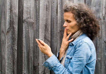 Profile view of mature woman in blue denim jacket using smart phone against grey wooden fence