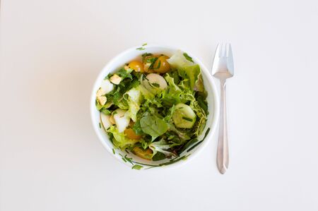 High angle view of salad with yellow tomatoes, cucumber, egg and lettuce leaves in a white bowl with a silver fork on a white tableの写真素材