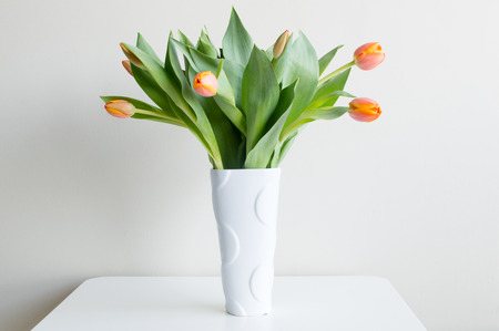 Orange tulips and green leaves in a white vase on a white table against a white wall