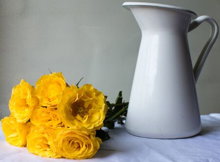 Yelllow roses laying next to white jug on a white tablecloth against a neutral backgroundの写真素材