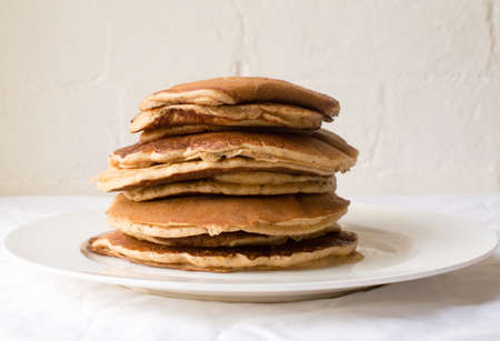 Stack of spelt flour pancakes on white plate on white tablecloth against white brick wall