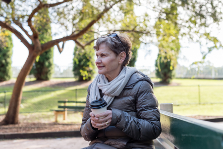 Older woman in scarf and coat sitting outdoors on park bench holding disposable coffee cup (selective focus)の写真素材