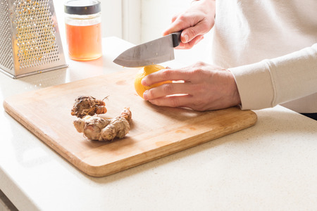 Middle aged woman cutting lemon on chopping board with turmeric, ginger root, honey and grater...