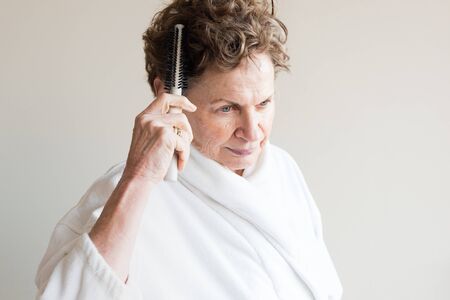 Head and shoulders view of older woman in white bathrobe brushing hairの写真素材